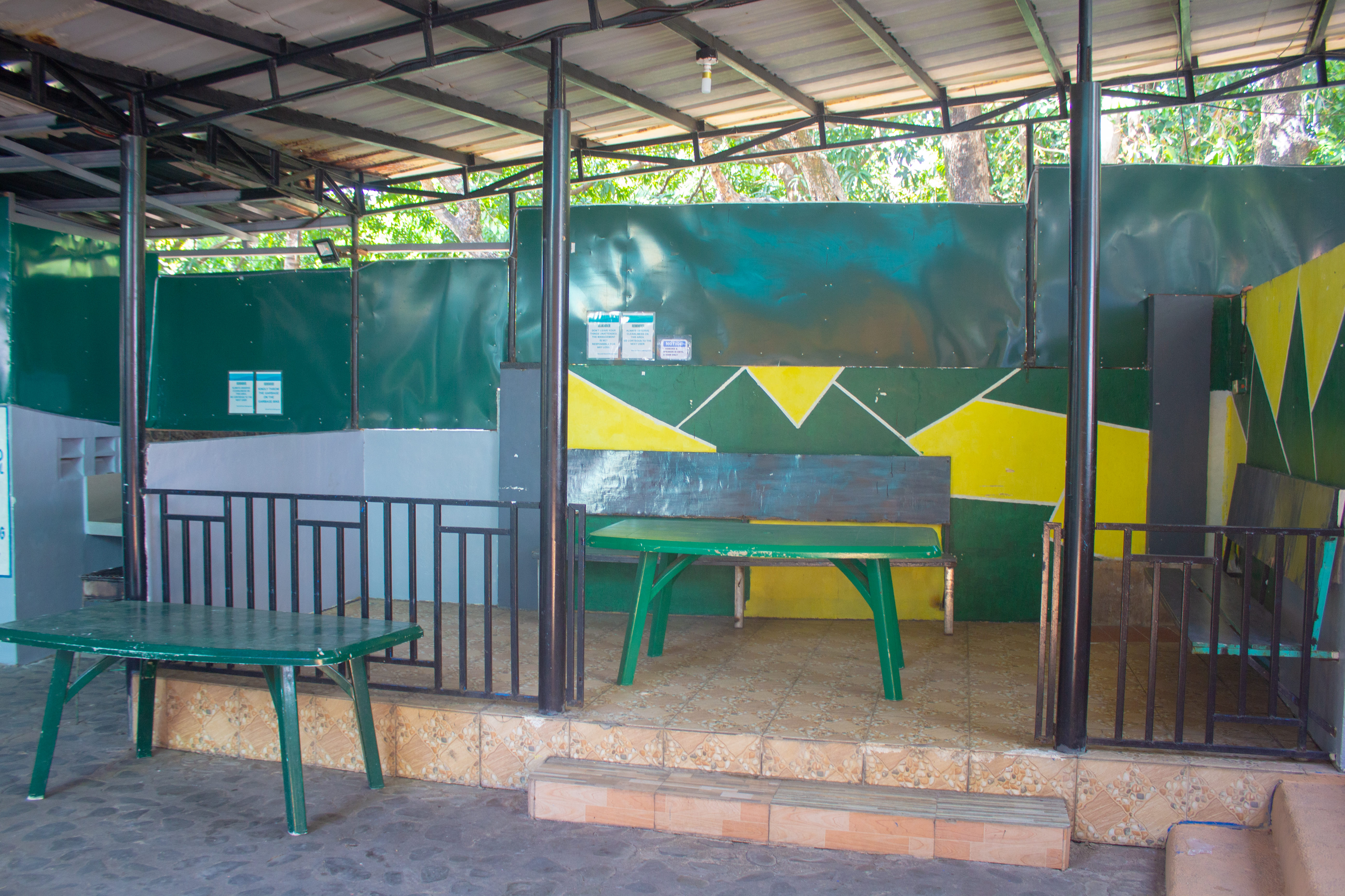 A small VIP seating area with a green and yellow geometric wall design, a raised tiled platform, metal railings, and green plastic tables and benches.